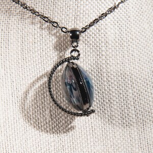 K&ouml;nnte beinhalten: Halskette mit silberfarbener Kette und Anh&auml;nger. Der Anh&auml;nger ist oval mit blau-wei&szlig;em Design in einem Metallrahmen. Kette und Rahmen haben eine dunkle, anthrazitfarbene Oberfl&auml;che. Die Halskette wird auf einer strukturierten, cremefarbenen Oberfl&auml;che pr&auml;sentiert.