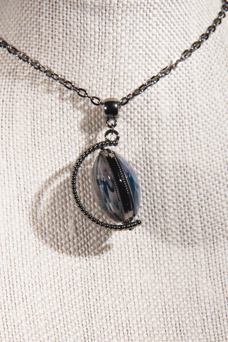 K&ouml;nnte beinhalten: Halskette mit silberfarbener Kette und Anh&auml;nger. Der Anh&auml;nger ist oval mit blau-wei&szlig;em Design in einem Metallrahmen. Kette und Rahmen haben eine dunkle, anthrazitfarbene Oberfl&auml;che. Die Halskette wird auf einer strukturierten, cremefarbenen Oberfl&auml;che pr&auml;sentiert.