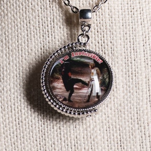 May include: Silver-toned pendant necklace featuring a circular pendant with a detailed image of two figures in a fight scene. The pendant has a beaded edge and the words "I'm Invincible!" are printed on the top. The chain is a delicate link style.