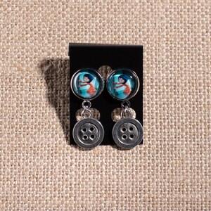 May include: A pair of silver dangle earrings with a blue and white button image and a silver button charm.