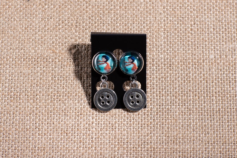 May include: A pair of silver dangle earrings with a blue and white button image and a silver button charm.