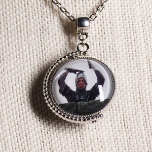 May include: Silver-toned necklace with a round pendant featuring a humorous image of a person in medieval attire. The pendant has a beaded edge and hangs from a delicate chain. A fun accessory for fans of comedy or historical themes.