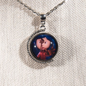 Puede incluir: Un collar de plata con un colgante redondo que presenta una imagen colorida de una ni&ntilde;a con cabello azul sosteniendo una llave. El colgante est&aacute; rodeado por un borde de cuentas plateadas.