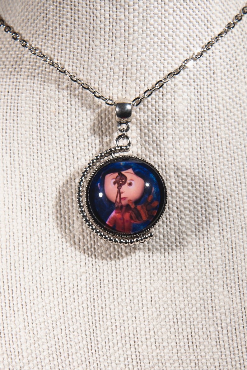 Puede incluir: Un collar de plata con un colgante redondo que presenta una imagen colorida de una ni&ntilde;a con cabello azul sosteniendo una llave. El colgante est&aacute; rodeado por un borde de cuentas plateadas.