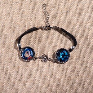 May include: Black suede bracelet featuring two round charms with images and a silver-tone flower accent. One charm depicts a cartoon character, the other a blue abstract design. Adjustable length.