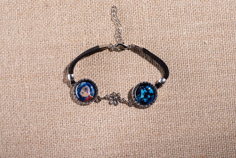 May include: Black suede bracelet featuring two round charms with images and a silver-tone flower accent. One charm depicts a cartoon character, the other a blue abstract design. Adjustable length.
