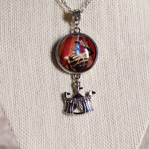 May include: Silver-toned necklace with a round pendant featuring a circus performer in a red and gold setting. Below the pendant hangs a detailed circus tent charm. The chain is delicate and the pendant is approximately 1 inch in diameter.