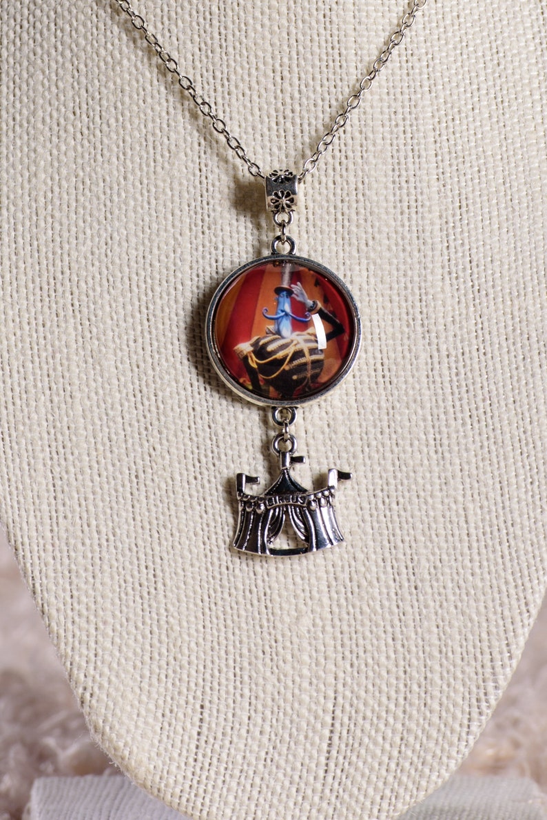 May include: Silver-toned necklace with a round pendant featuring a circus performer in a red and gold setting. Below the pendant hangs a detailed circus tent charm. The chain is delicate and the pendant is approximately 1 inch in diameter.