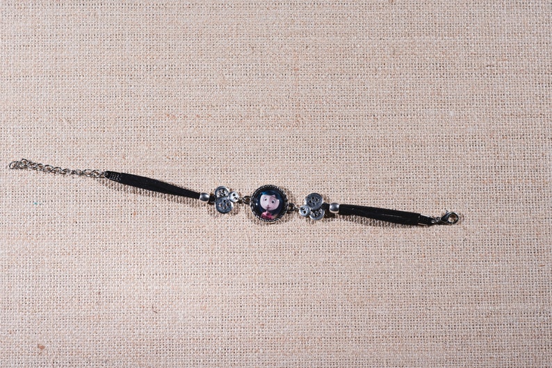 Peut inclure: Un bracelet en cuir noir avec une cha&icirc;ne argent&eacute;e et un charme rond repr&eacute;sentant une fille de dessin anim&eacute;. Le charme est entour&eacute; de deux charmes argent&eacute;s &agrave; motif floral.