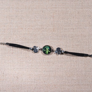 Peut inclure: Un bracelet en cuir noir avec une cha&icirc;ne argent&eacute;e et un cam&eacute;e rond vert repr&eacute;sentant une figure stylis&eacute;e. Le bracelet est orn&eacute; d'accents argent&eacute;s.