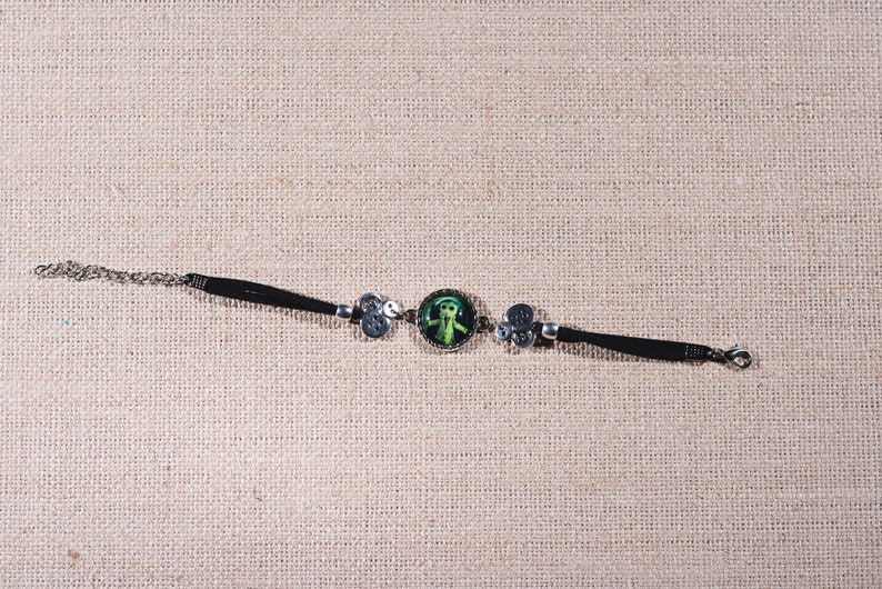 Peut inclure: Un bracelet en cuir noir avec une cha&icirc;ne argent&eacute;e et un cam&eacute;e rond vert repr&eacute;sentant une figure stylis&eacute;e. Le bracelet est orn&eacute; d'accents argent&eacute;s.