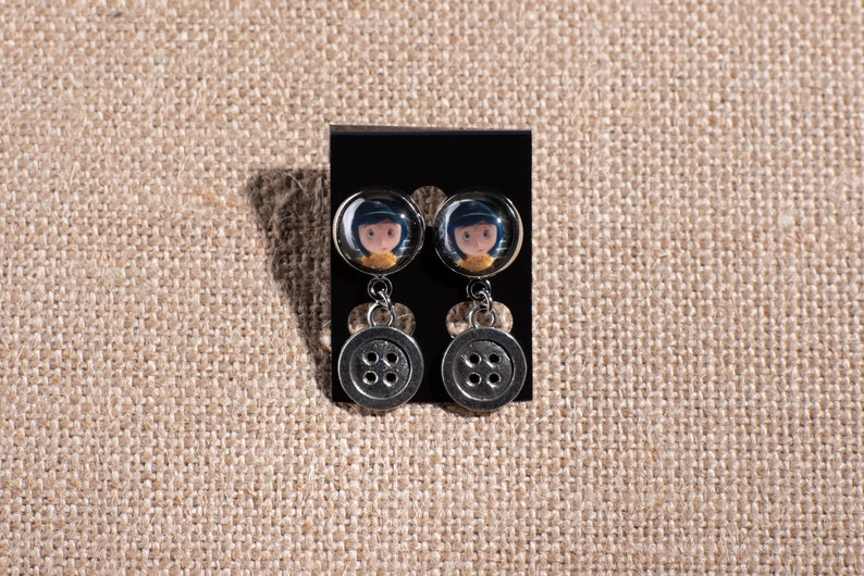 May include: A pair of dangle earrings with a button charm. The top of the earring features a round glass cabochon with a cartoon image of a girl with black hair and a yellow shirt.