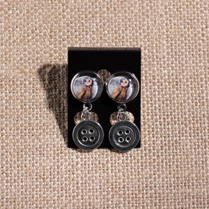 May include: A pair of silver dangle earrings with a round button charm and a smaller round charm featuring a girl in a brown coat.