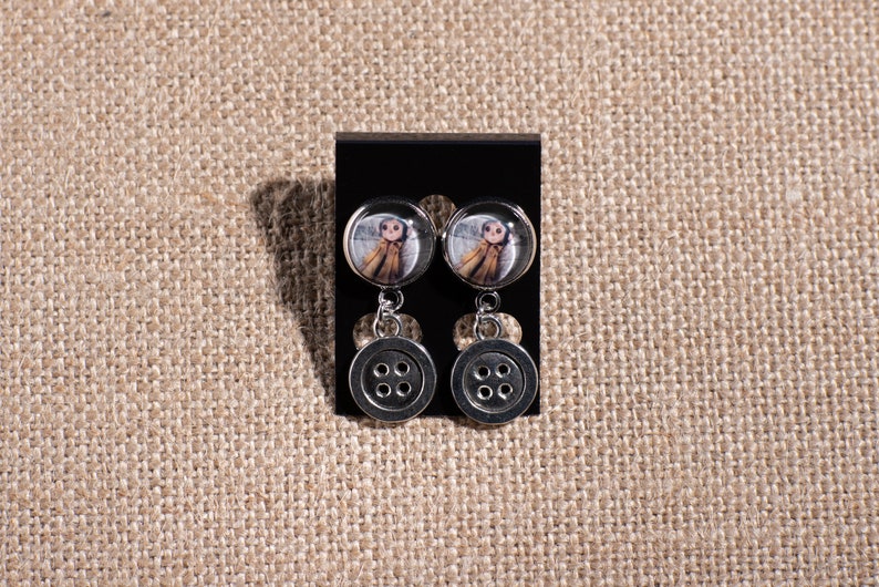 May include: A pair of silver dangle earrings with a round button charm and a smaller round charm featuring a girl in a brown coat.