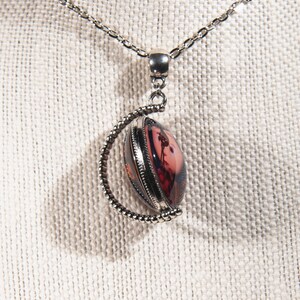Puede incluir: Un collar de cadena de plata con un colgante redondo que presenta un dise&ntilde;o floral rosa y rojo. El colgante est&aacute; enmarcado en plata y tiene un borde de cuentas.