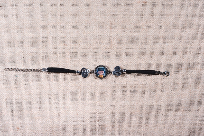 Peut inclure: Un bracelet en cuir noir avec une cha&icirc;ne argent&eacute;e et un charme repr&eacute;sentant une fille de dessin anim&eacute;. Le charme est rond et a une bordure argent&eacute;e.