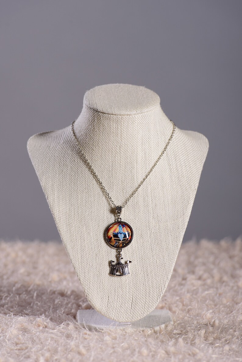 May include: Silver-toned necklace with a pendant featuring a colorful, circular design and a small, decorative charm. The pendant has a detailed image with blue, orange, and yellow hues. The necklace is displayed on a white mannequin.