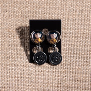 May include: A pair of silver dangle earrings with a black button charm and a round glass charm featuring a girl with brown hair and a yellow jacket.