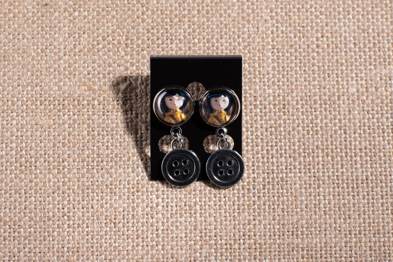 May include: A pair of silver dangle earrings with a black button charm and a round glass charm featuring a girl with brown hair and a yellow jacket.