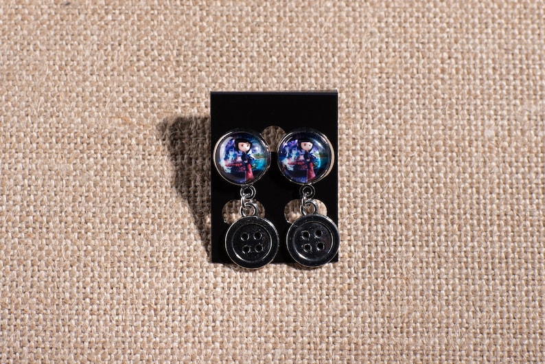 May include: A pair of dangle earrings with a black button charm and a round glass charm featuring a colorful illustration of a girl.
