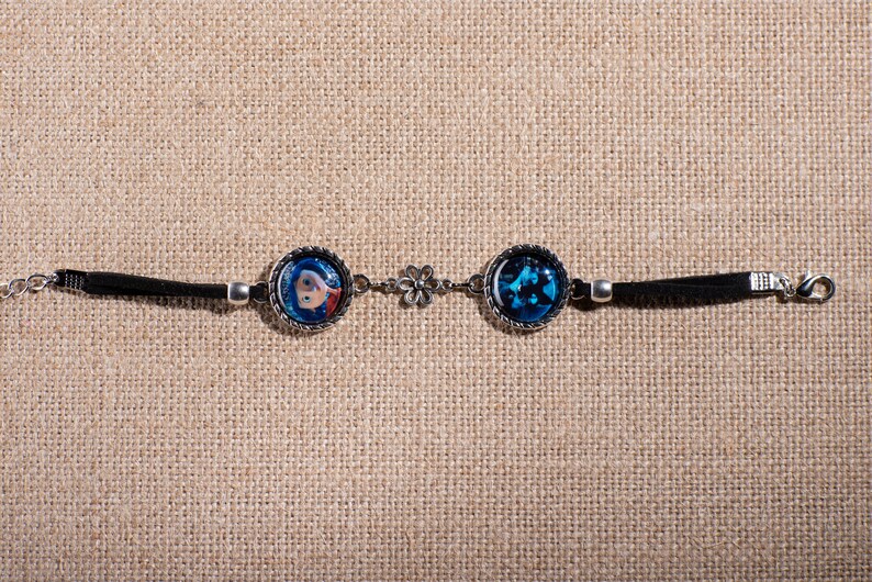 May include: A silver-toned bracelet featuring two circular charms with images. One charm displays a blue-haired character, the other a blue, abstract design. The bracelet has a black cord and a flower-shaped connector.