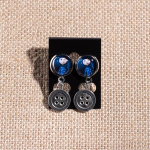 May include: A pair of silver dangle earrings with a blue and white image of a girl on the top and a silver button on the bottom.