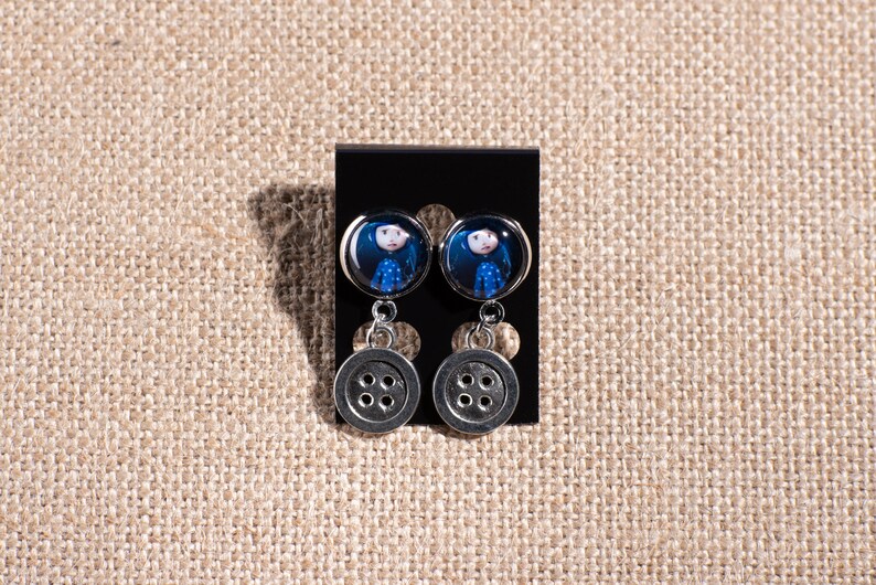 May include: A pair of silver dangle earrings with a blue and white image of a girl on the top and a silver button on the bottom.