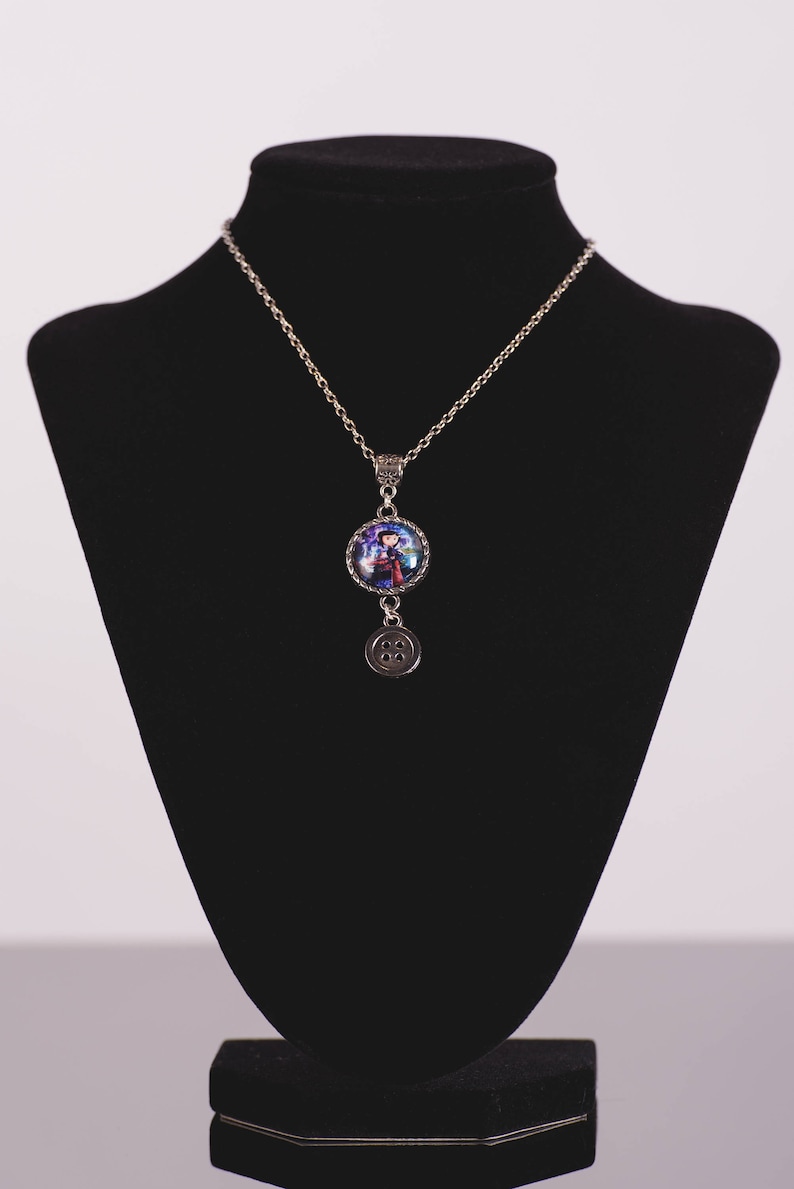 May include: Silver-toned necklace with a round pendant featuring a colorful image of a character. A smaller, circular charm with a button design hangs below the pendant. The necklace is displayed on a black mannequin.