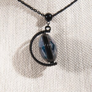 K&ouml;nnte beinhalten: Schwarze Halskette mit einem Anh&auml;nger mit einem schwarzen Rahmen um ein blaues und wei&szlig;es Design. Der Anh&auml;nger h&auml;ngt an einer schwarzen Kette mit einer kleinen schwarzen Perle. Die Halskette wird auf einem neutralfarbenen Stoff pr&auml;sentiert.