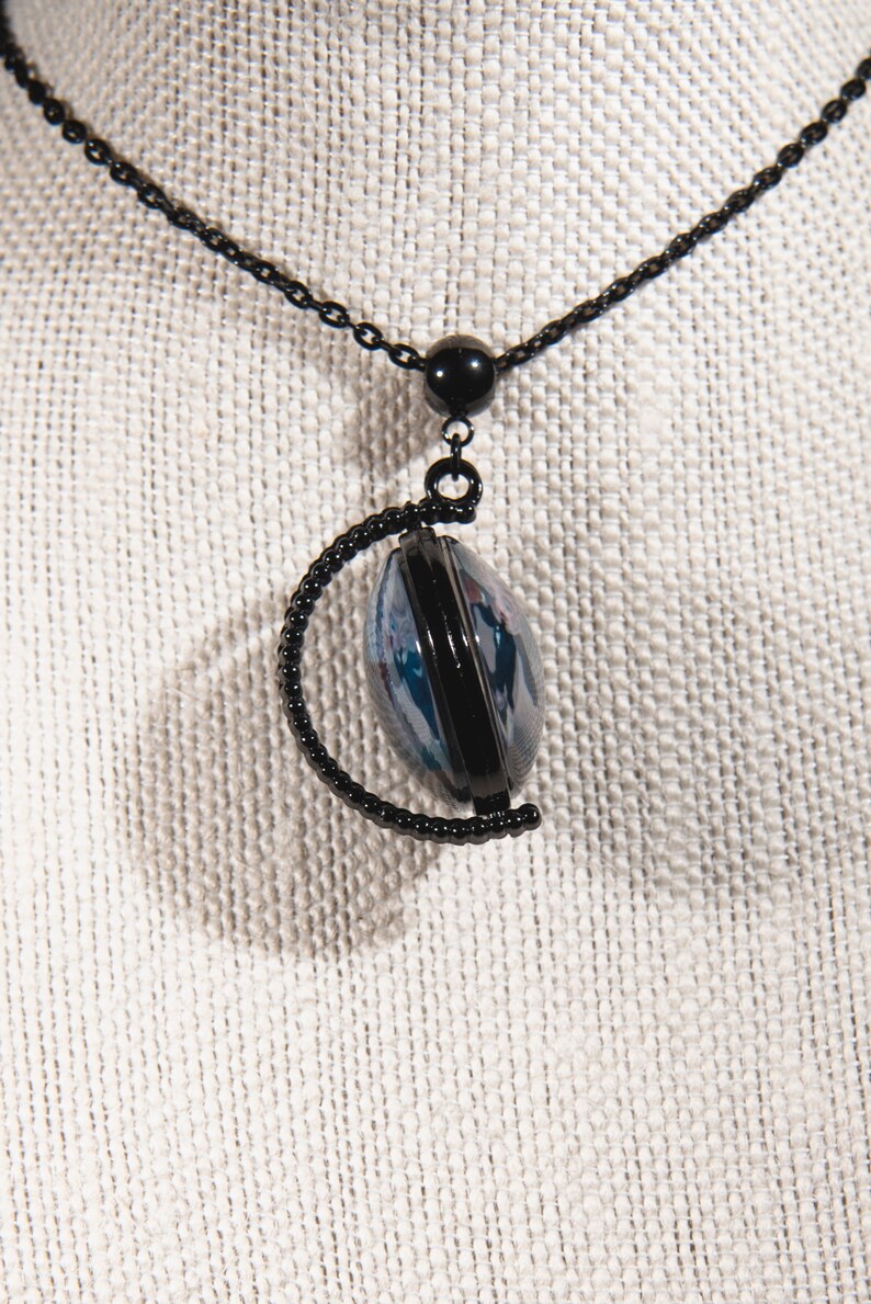 K&ouml;nnte beinhalten: Schwarze Halskette mit einem Anh&auml;nger mit einem schwarzen Rahmen um ein blaues und wei&szlig;es Design. Der Anh&auml;nger h&auml;ngt an einer schwarzen Kette mit einer kleinen schwarzen Perle. Die Halskette wird auf einem neutralfarbenen Stoff pr&auml;sentiert.