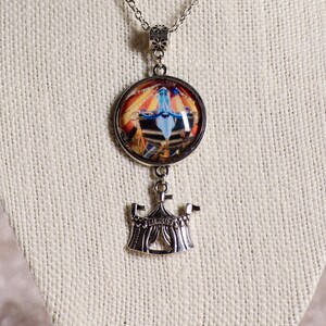 May include: Silver-toned necklace featuring a round pendant with a circus scene and a smaller circus tent charm. The pendant showcases a colorful circus performance with a blue figure. The tent charm has the word "CIRCUS" on it.