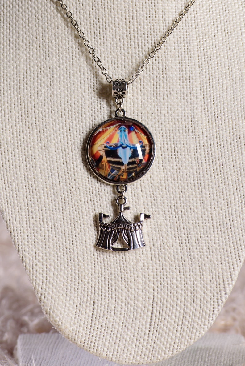 May include: Silver-toned necklace featuring a round pendant with a circus scene and a smaller circus tent charm. The pendant showcases a colorful circus performance with a blue figure. The tent charm has the word "CIRCUS" on it.