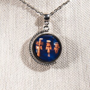 Puede incluir: Un collar de cadena de plata con un colgante redondo con fondo azul con tres &aacute;ngeles en blanco y naranja. El colgante est&aacute; rodeado por un borde de cuentas plateadas.