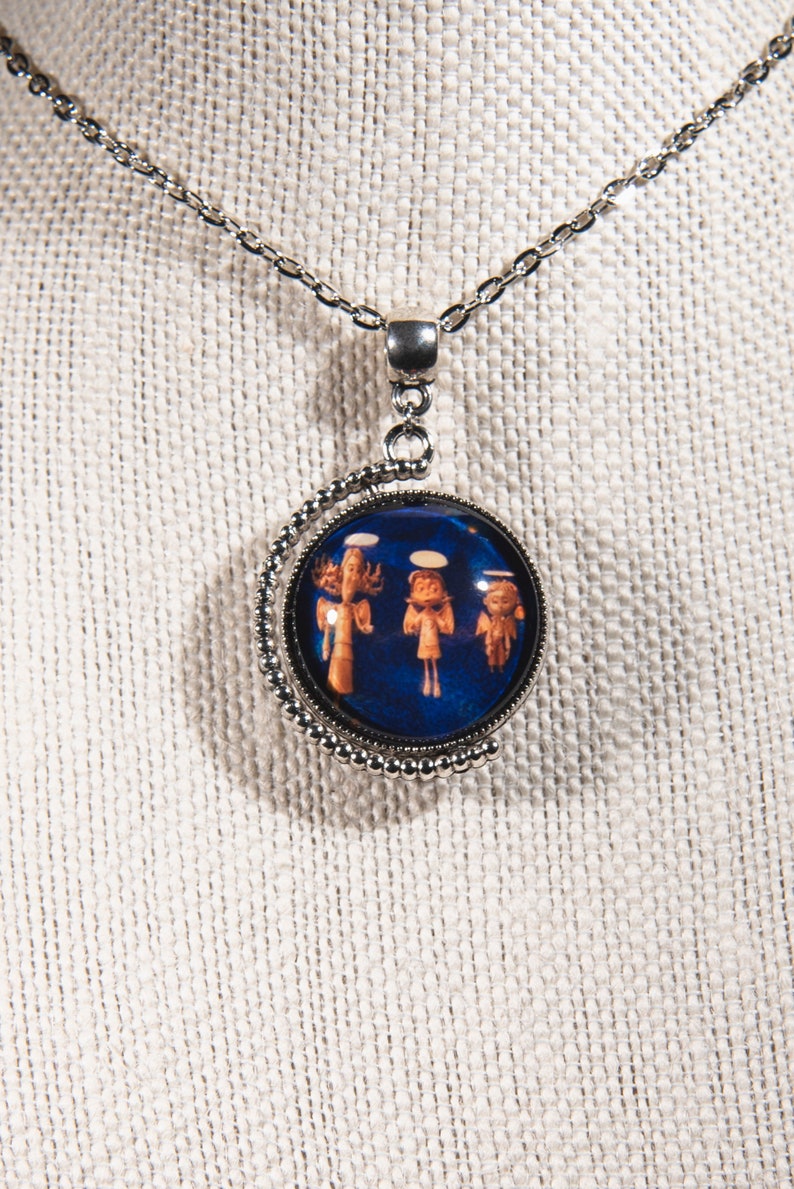 Puede incluir: Un collar de cadena de plata con un colgante redondo con fondo azul con tres &aacute;ngeles en blanco y naranja. El colgante est&aacute; rodeado por un borde de cuentas plateadas.