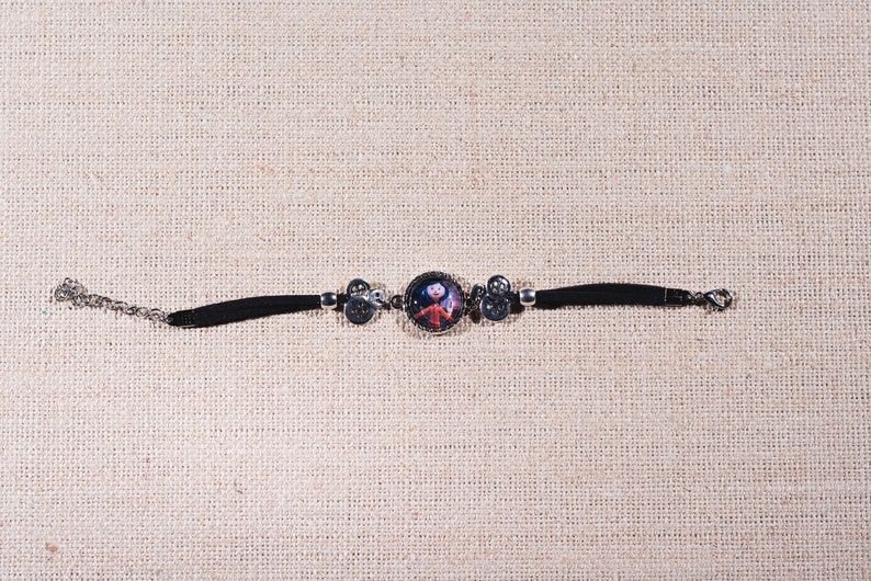 Peut inclure: Un bracelet en cuir noir avec un charme argent&eacute; repr&eacute;sentant un personnage de dessin anim&eacute;. Le charme est rond et pr&eacute;sente une image color&eacute;e d'une fille avec une robe rouge et un fond bleu.