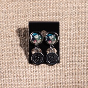 May include: A pair of dangle earrings with a black button charm and a round glass charm featuring a cartoon girl with blue hair and a green dress.