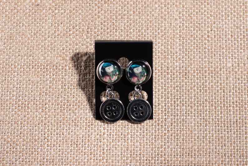 May include: A pair of dangle earrings with a black button charm and a round glass charm featuring a cartoon girl with blue hair and a green dress.