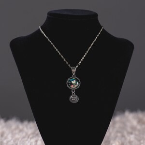 May include: Silver-toned necklace with a round pendant featuring a colorful image. Below the pendant hangs a charm with a button design. The necklace is displayed on a black jewelry bust against a neutral background.