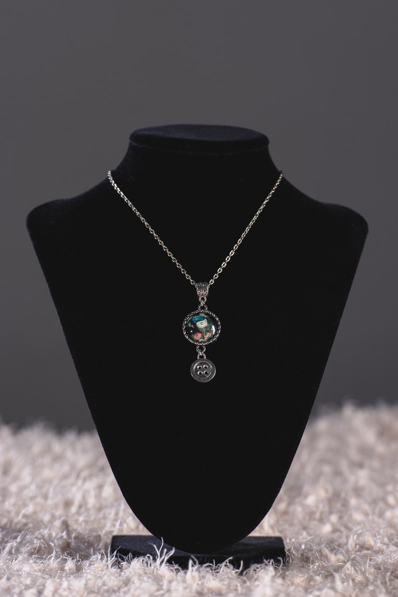 May include: Silver-toned necklace with a round pendant featuring a colorful image. Below the pendant hangs a charm with a button design. The necklace is displayed on a black jewelry bust against a neutral background.