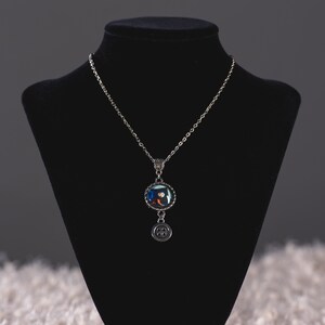 May include: A silver-toned necklace with a round pendant featuring a colorful design. The pendant is connected to a chain and has a small button charm hanging below. The necklace is displayed on a black bust.