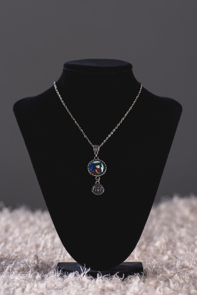 May include: A silver-toned necklace with a round pendant featuring a colorful design. The pendant is connected to a chain and has a small button charm hanging below. The necklace is displayed on a black bust.