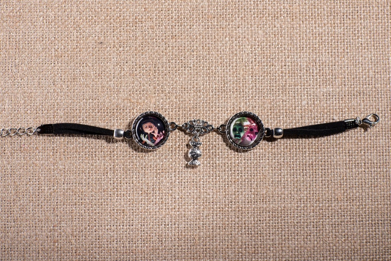 May include: A black leather bracelet with two silver round charms featuring colorful images. The charms are connected by a silver chain with a dangling silver charm.