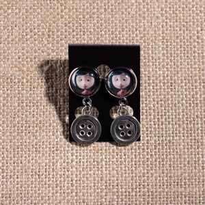 May include: A pair of silver dangle earrings with a black button charm and a round glass charm featuring a character from the movie "Coraline".