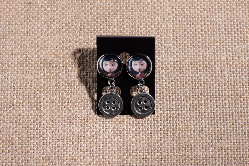 May include: A pair of silver dangle earrings with a black button charm and a round glass charm featuring a character from the movie "Coraline".