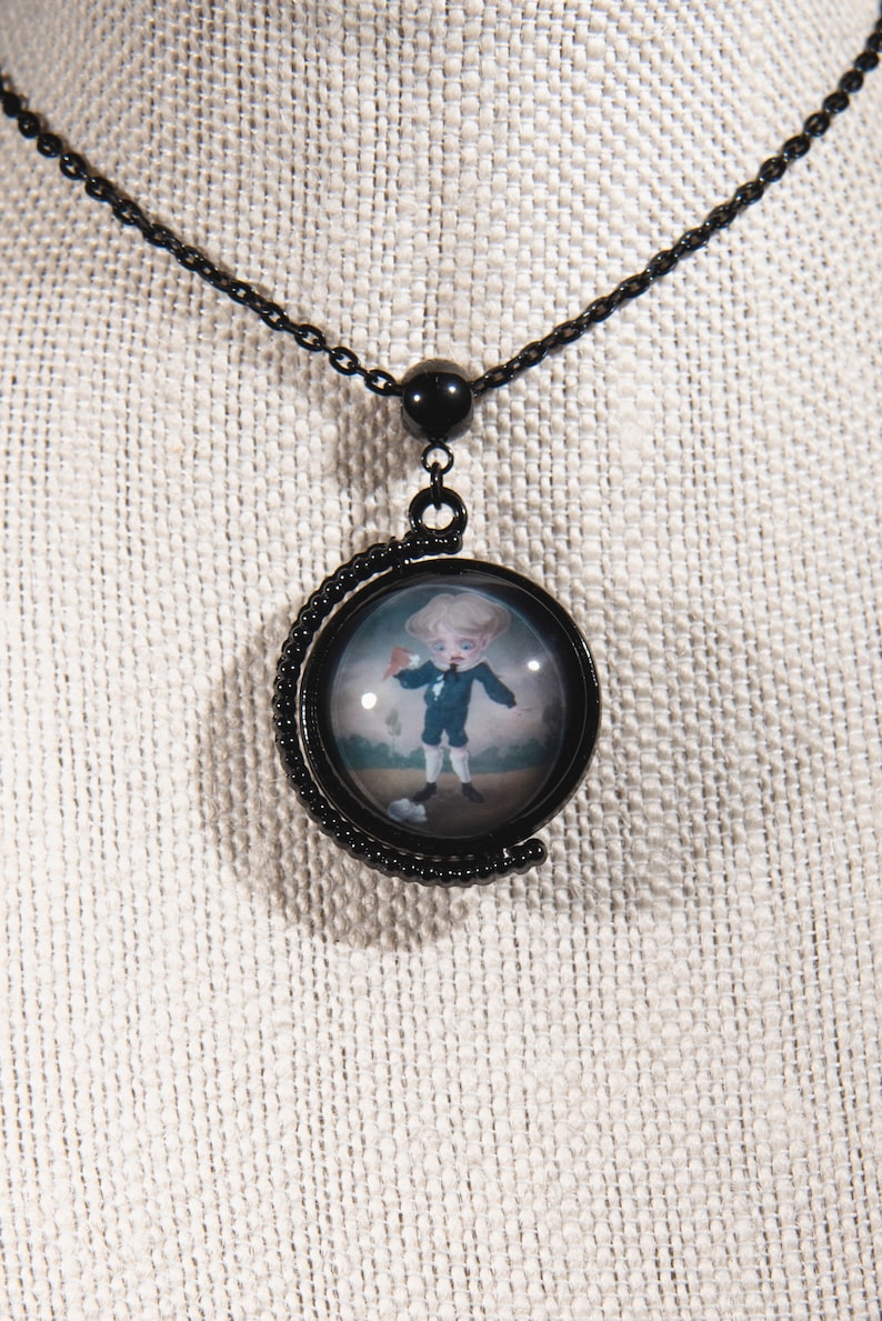 K&ouml;nnte beinhalten: Schwarze Metall-Anh&auml;nger-Halskette mit einem runden Anh&auml;nger mit einem Miniaturgem&auml;lde eines Jungen in einem blauen Anzug. Der Anh&auml;nger h&auml;ngt an einer schwarzen Kette. Der Junge h&auml;lt einen roten Gegenstand.