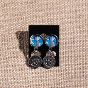 May include: A pair of silver dangle earrings with a blue and white image of a girl with a red jacket and a button charm.