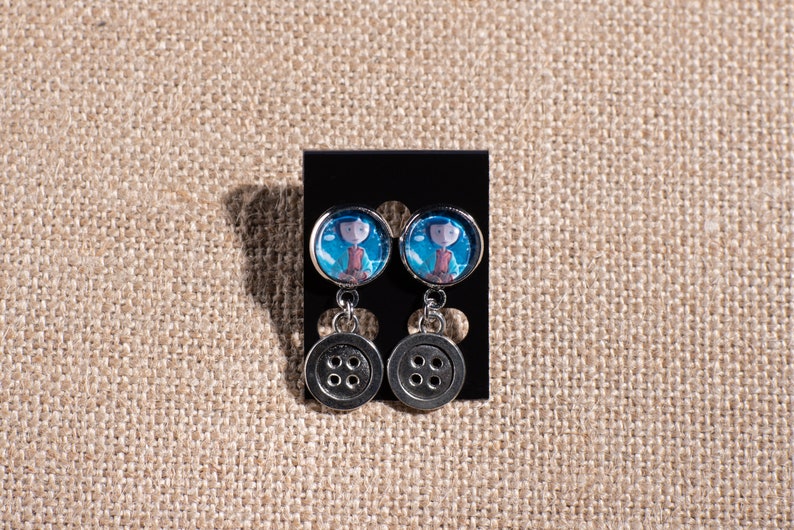 May include: A pair of silver dangle earrings with a blue and white image of a girl with a red jacket and a button charm.