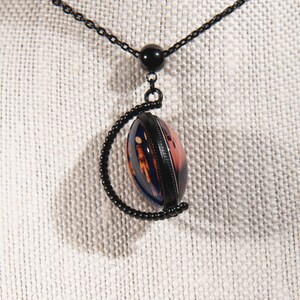 Puede incluir: Un collar de cadena negra con un colgante redondo que presenta un dise&ntilde;o abstracto colorido encerrado en un marco de metal negro. El colgante est&aacute; unido a la cadena con una cuenta negra.