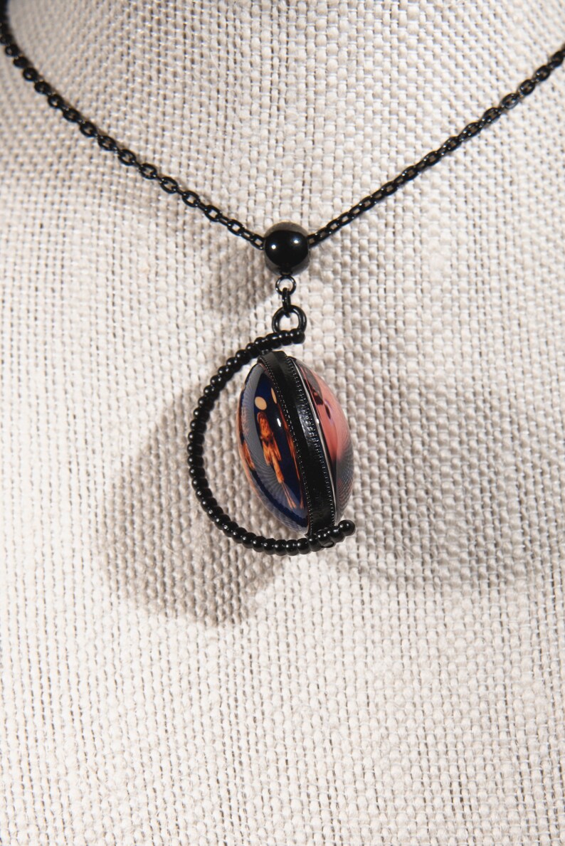 Puede incluir: Un collar de cadena negra con un colgante redondo que presenta un dise&ntilde;o abstracto colorido encerrado en un marco de metal negro. El colgante est&aacute; unido a la cadena con una cuenta negra.