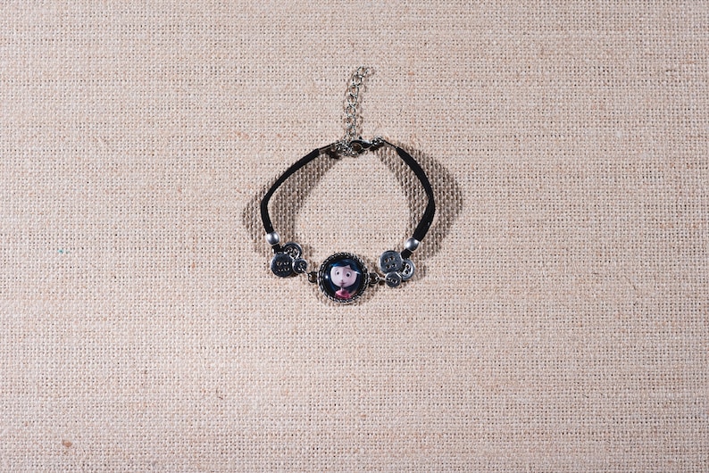 Peut inclure: Un bracelet noir avec un charme argent&eacute; repr&eacute;sentant une fille de dessin anim&eacute; aux cheveux noirs et une robe rose. Le charme est entour&eacute; de plus petits charmes argent&eacute;s.